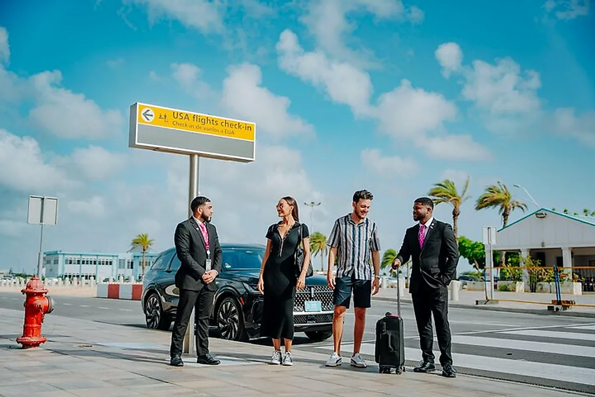 Two suited drivers greeting arriving travellers outside Aruba's airport terminal, beside a waiting SUV