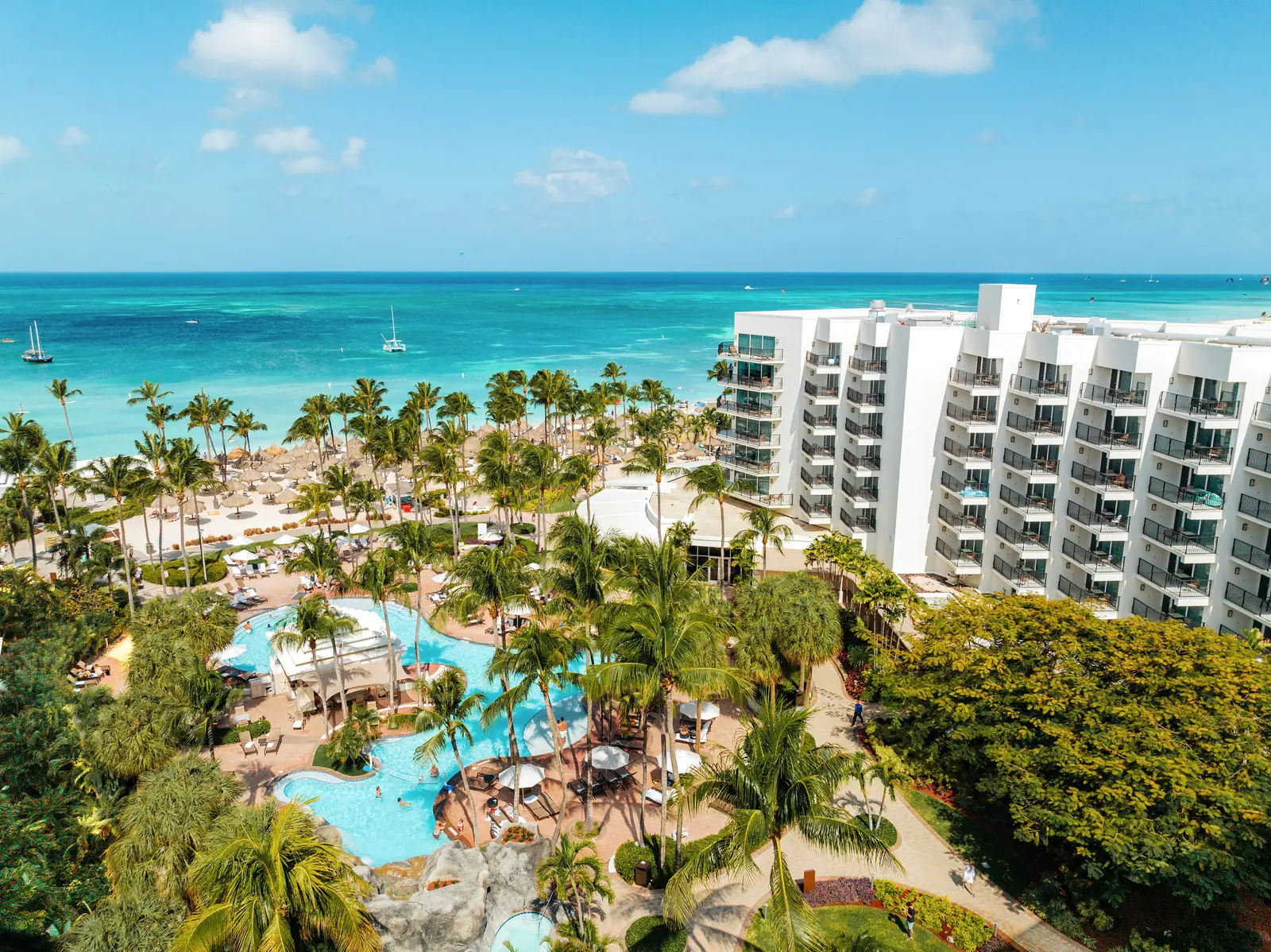 Aerial view of a Palm Beach resort in Aruba — pool and palms in the foreground, turquoise water and catamarans beyond